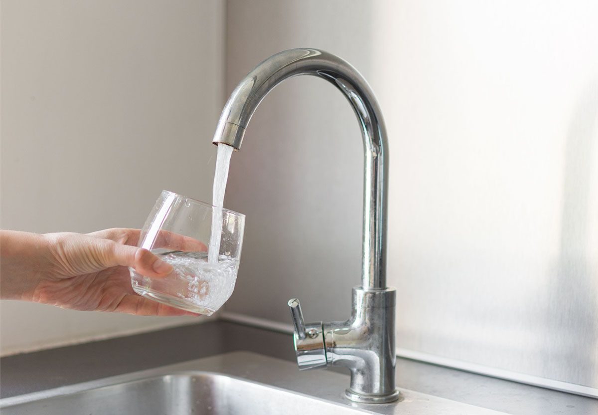 A hand holds a glass under a modern kitchen faucet, filling it with running water above a stainless steel sink.