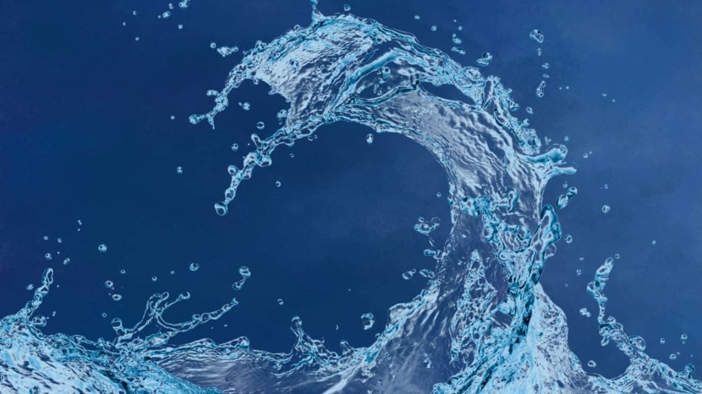 A splash of clear blue water forms the shape of a curling wave or arch against a dark blue background, with droplets scattering in various directions.