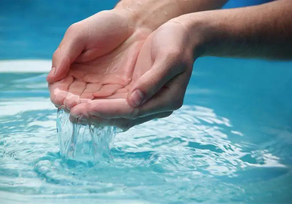 A pair of cupped hands holds clear water above a pool, with water gently flowing through the fingers back into the pool below.