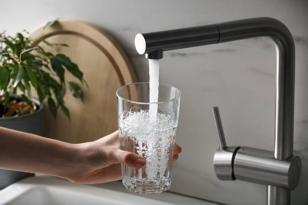 A hand holds a clear glass under a modern kitchen faucet as water pours into the glass, creating bubbles. A plant and a round wooden cutting board are visible in the background.
