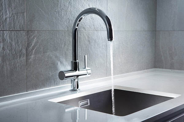 A modern chrome kitchen faucet with water running into a stainless steel sink, set against a gray tiled backsplash and a white countertop.