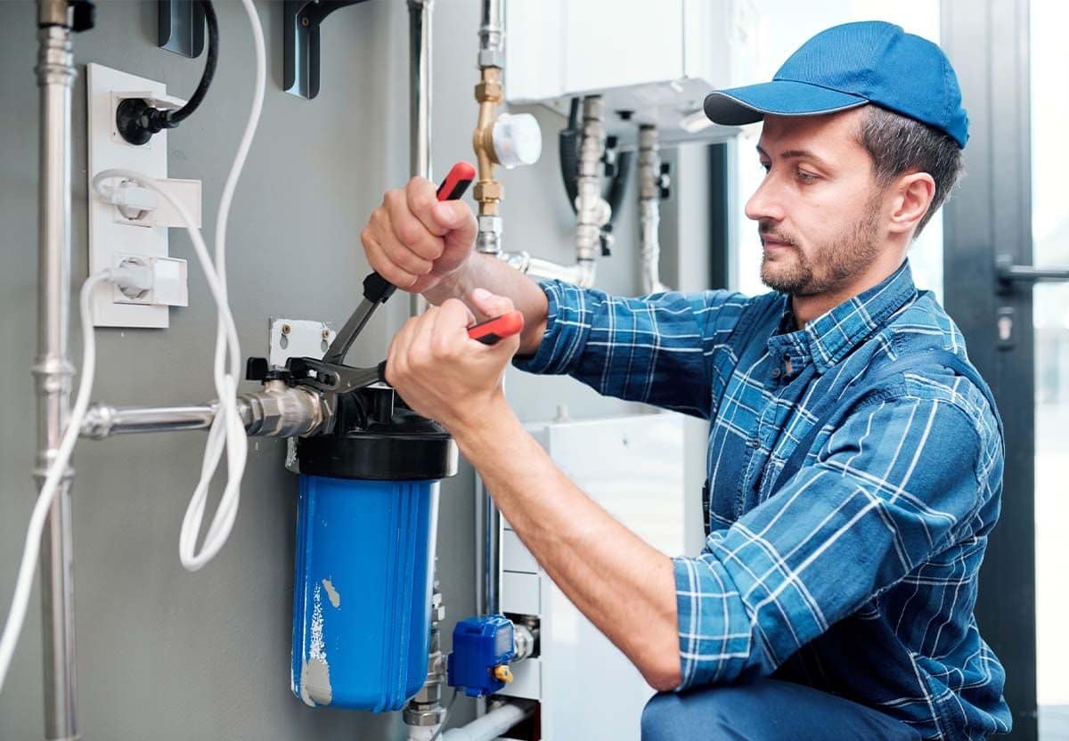 A man wearing a blue cap and plaid shirt uses a wrench to work on a blue water filter attached to pipes, likely performing maintenance or installation in an indoor utility area.