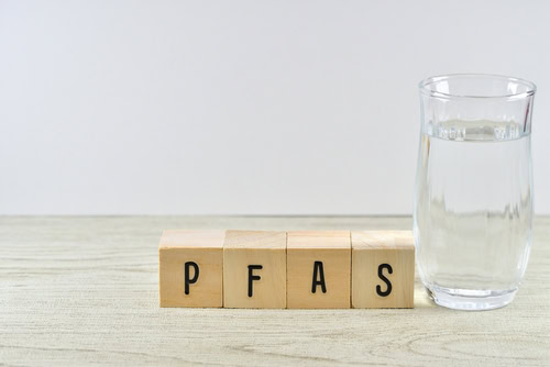 A glass of water sits on a wooden surface next to wooden blocks spelling out "PFAS" in black letters, against a plain light background.