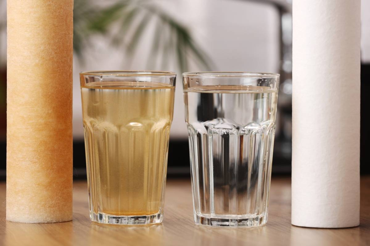 Two glasses of water sit side by side; the left glass contains yellowish, murky water, while the right glass contains clear water. Two cylindrical water filters stand upright on either side of the glasses.