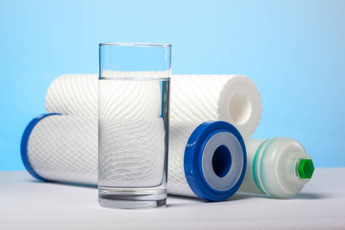 A clear glass of water stands in front of three white water filter cartridges with blue and green ends, set against a light blue background.