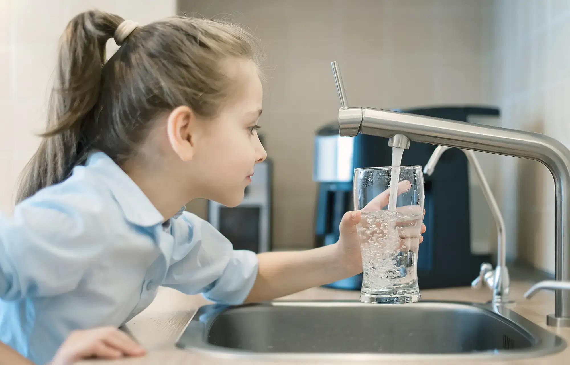 A young girl with a ponytail fills a clear glass with water from a modern kitchen faucet, standing by the sink and watching the water flow.