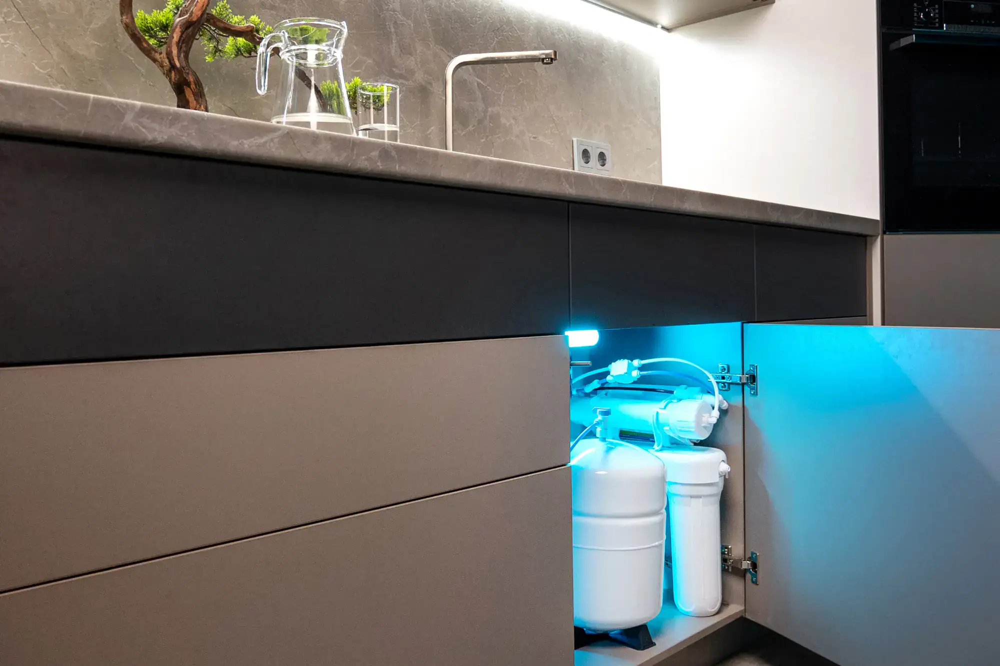 A modern kitchen with a marble countertop and an open cabinet under the sink, revealing a water filtration system with tanks and filters illuminated by a blue light.