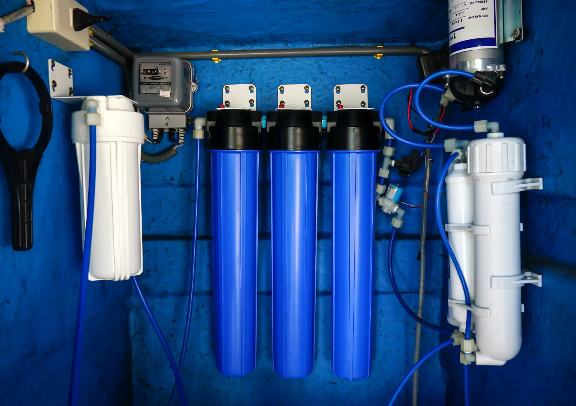 A water filtration system with three large blue filter housings, white filter units, pipes, cables, and fittings mounted on a blue wall.