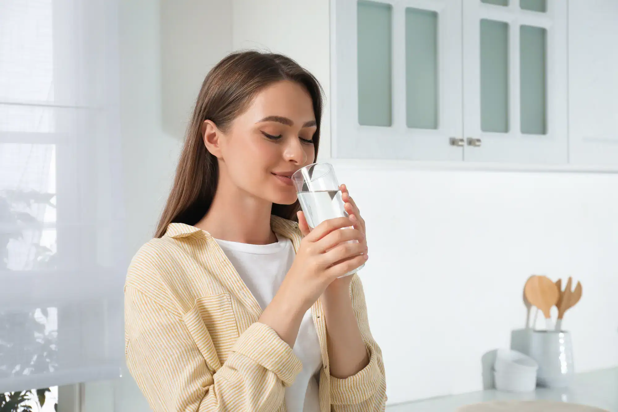 A young woman with long brown hair, wearing a yellow striped shirt, stands in a bright kitchen, smiling with her eyes closed as she holds and drinks a glass of water.