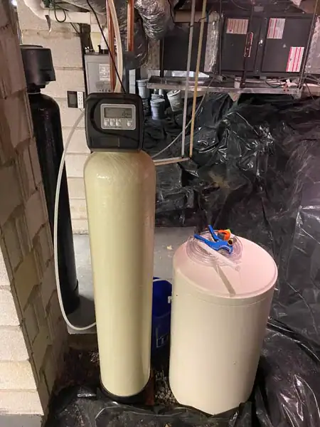 A water softener system in a basement, featuring a tall beige tank with a digital control head and a round white salt brine tank beside it, surrounded by exposed pipes and black plastic sheeting on the floor.